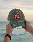WWJD camo hat held against an ocean sunset with the red-and-white front design visible