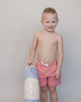 Young boy in red checkered swim trunks holding a colorful beach ball against a plain background