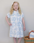 Young girl wearing a blue floral dress standing next to a woven basket with towels on a plain background