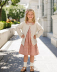 Young girl in a dress standing on a sunlit sidewalk with a building and trees in the background