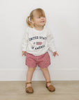 Child wearing a 'United States of America' sweatshirt and red checkered shorts on a white background