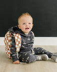 Baby sitting on a wooden floor wearing a checkered backpack against a black wall.