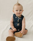 Baby sitting on a white blanket wearing a navy blue sleeveless dress with buttons and brown shoes.