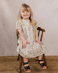 Young girl in a floral dress sitting on a wooden chair against a plain background