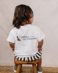 Child wearing a white t-shirt with 'Mebie Outdoors' logo, sitting on a wooden stool against a beige wall.