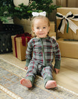 Child in plaid pajamas sitting on the floor with a Christmas tree and presents in the background