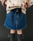Person wearing a patterned cardigan, denim skirt, gray tights, and white shoes on a black background