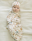 Newborn baby swaddled in a patterned blanket with a matching hat on a soft beige surface