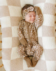 Newborn baby in a brown patterned outfit and headband lying on a checkered blanket in a wooden crib.