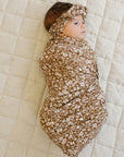 Baby wrapped in a floral swaddle and headband on a beige quilted surface