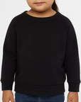 Toddler girl wearing a black BELLA + CANVAS 3901T sponge fleece raglan crewneck sweatshirt, front view, neutral background