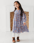 Girl wearing a blue floral long sleeve flared dress with white tights and brown boots beside a white fireplace.