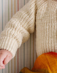 Child wearing a beige knitted sweater with colorful pants against a striped background