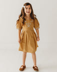 Girl wearing a mustard dress with floral embroidery, short puff sleeves, and brown shoes.
