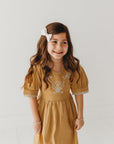 Front view of a girl wearing the mustard dress, showing the embroidered floral yoke and gathered skirt.