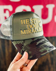 Camouflage cap with 'He Still Does Miracles' text held by a hand, with a blurred background.