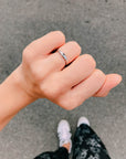 Silver-tone cross promise ring worn on a hand against a grey pavement background