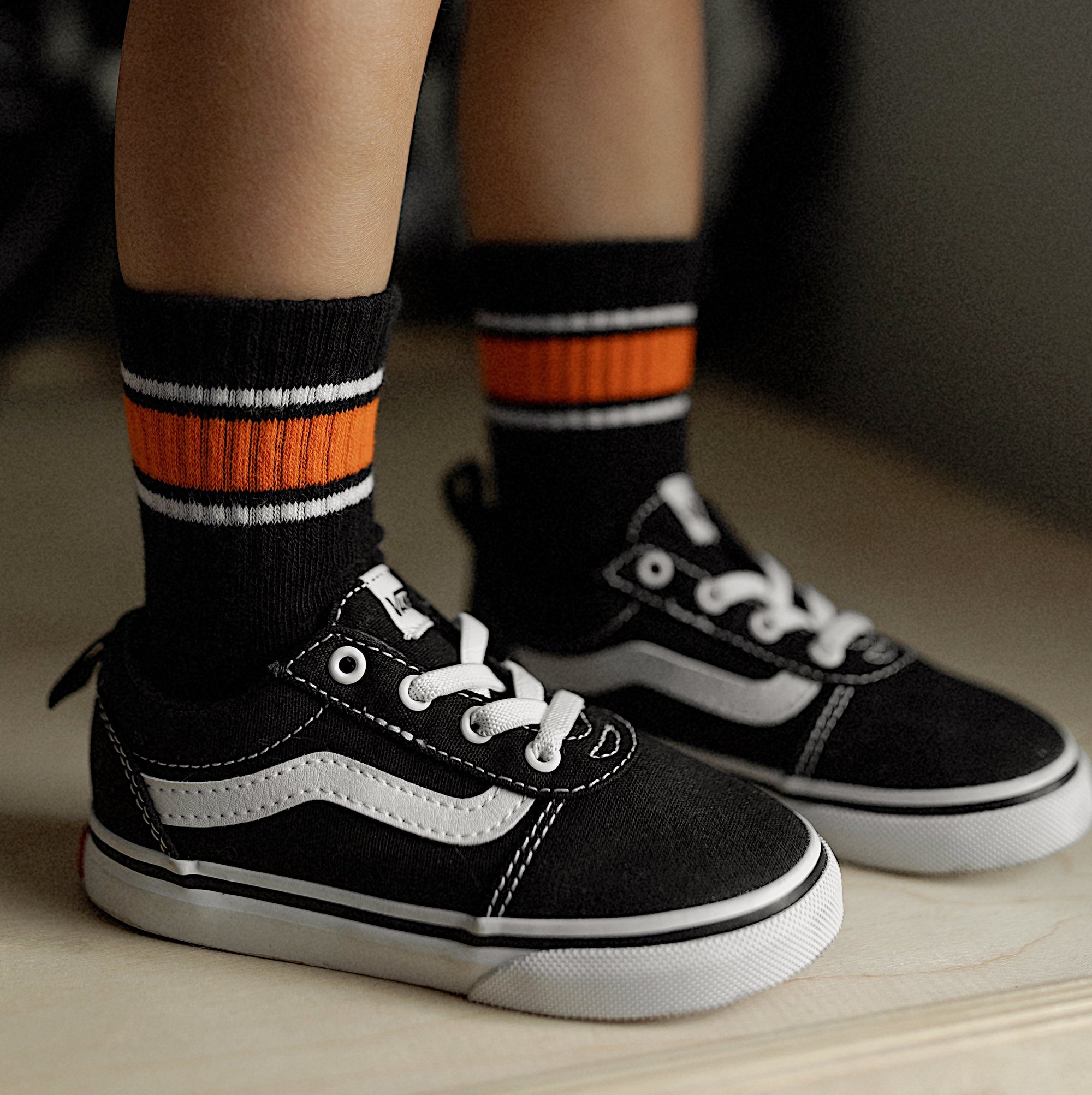 Child's legs wearing black sneakers with white laces, black and orange socks, and checkered shorts.