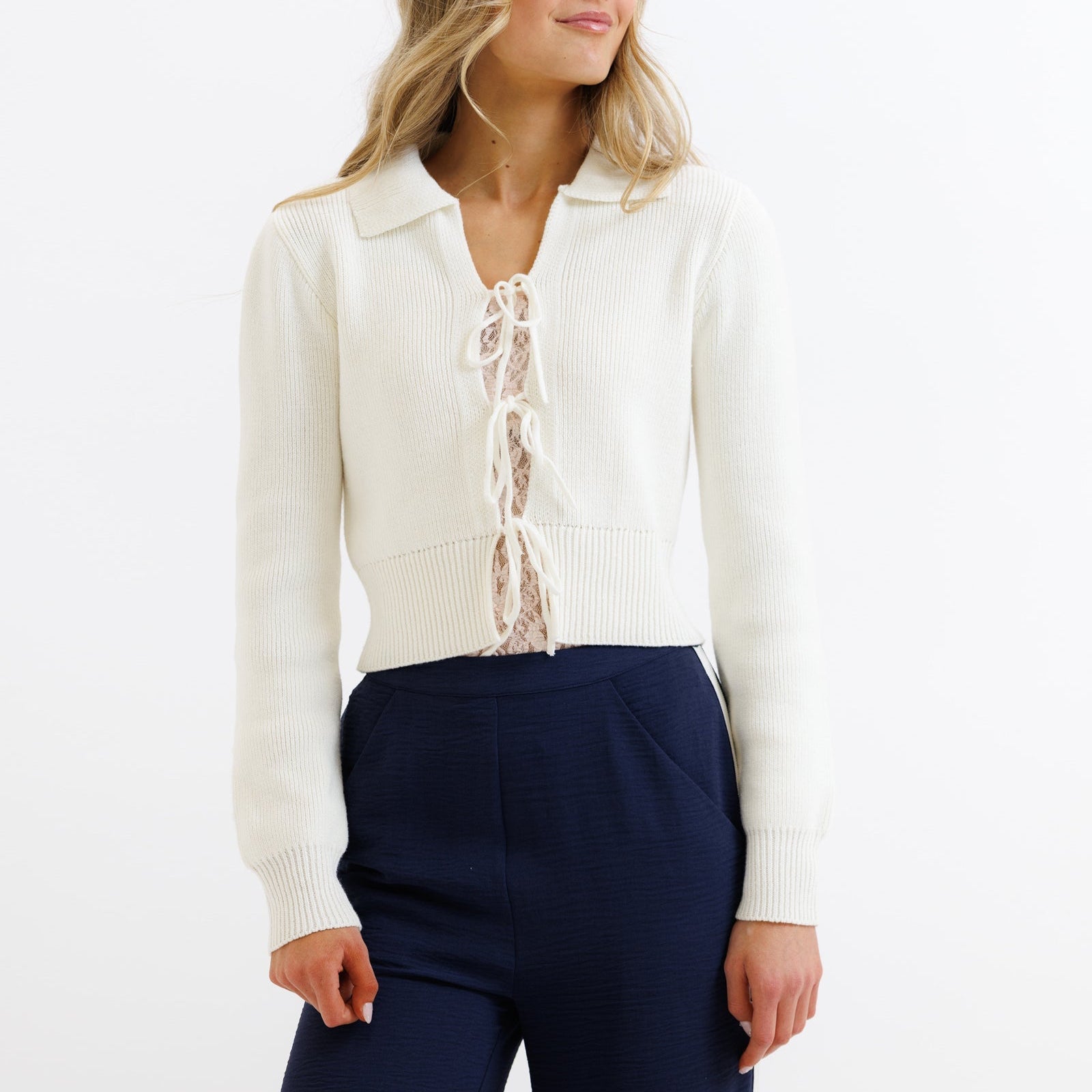 Woman wearing a white cardigan over a lace top with navy pants on a white background