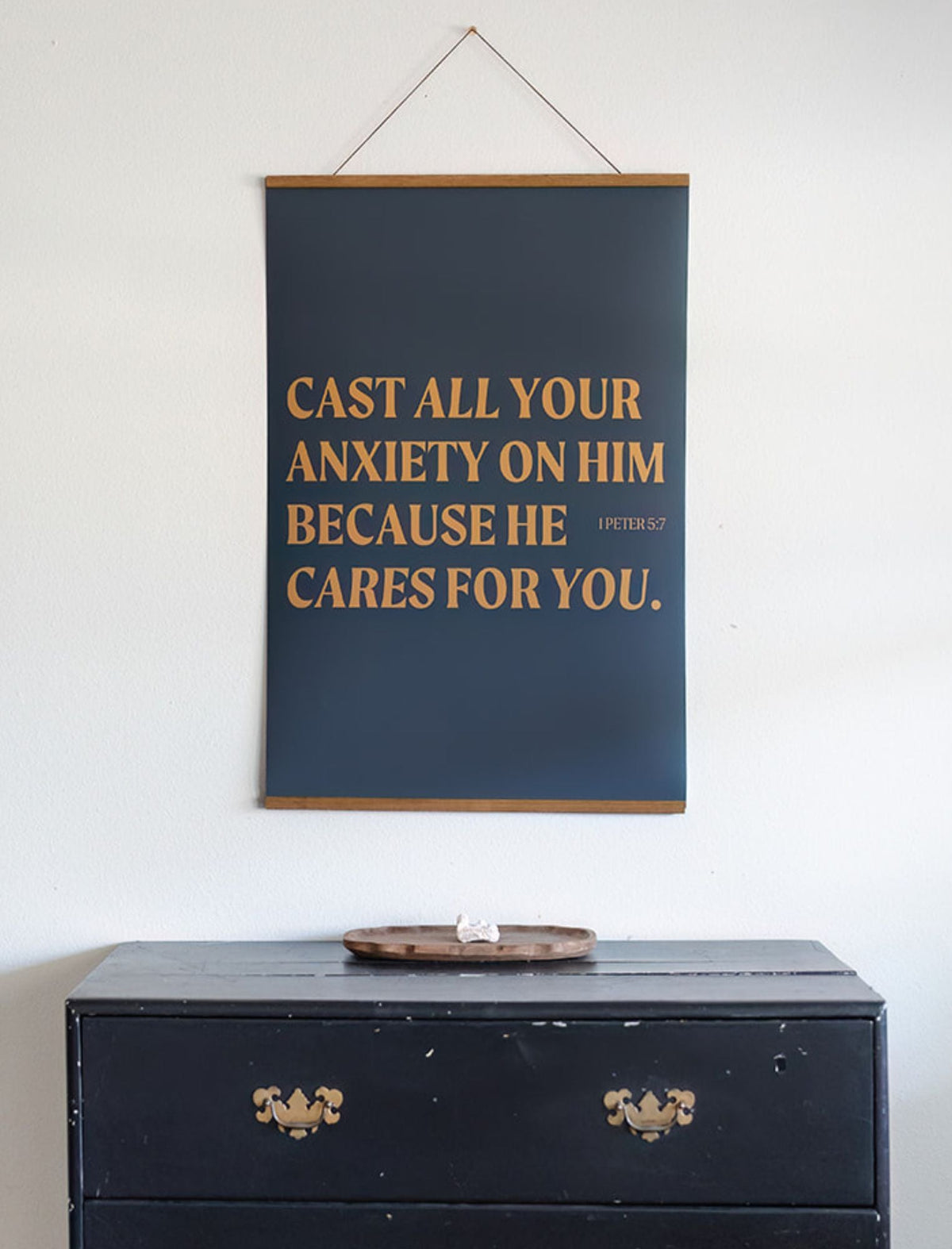 Deep blue Cast All Your Anxiety scripture poster with golden lettering and wooden hanging rails displayed above a dark cabinet.
