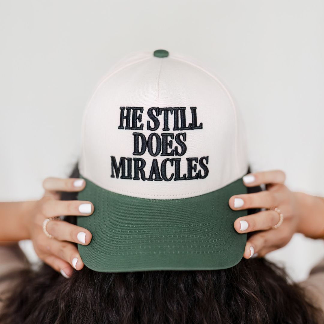 Close-up of He Still Does Miracles embroidery on a cream and green baseball cap