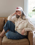 Woman seated in an armchair wearing a Jireh embroidered hat with a cream jacket and jeans