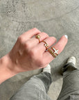 White enamel cross gold-tone ring styled with other rings on a hand above a gray textured surface