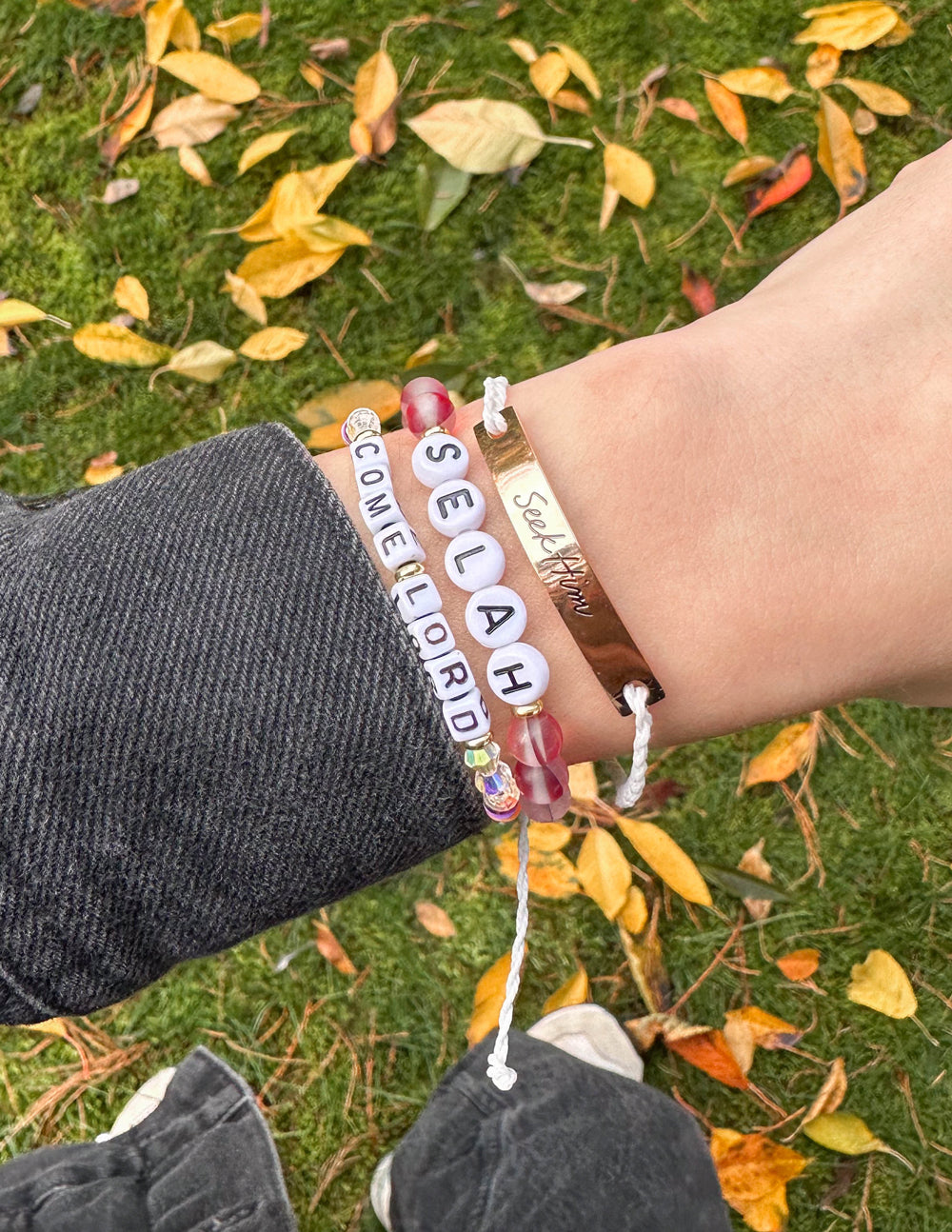 Seek Him cream string bracelet layered with other bracelets on a wrist above autumn leaves
