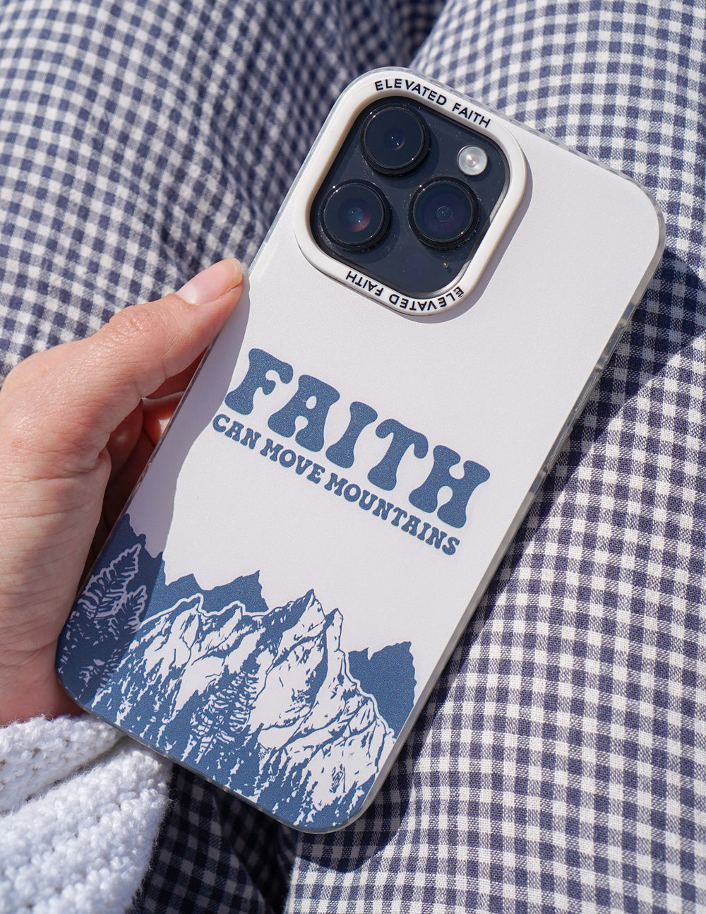 Hand holding the Faith Can Move Mountains iPhone case against blue and white checked fabric