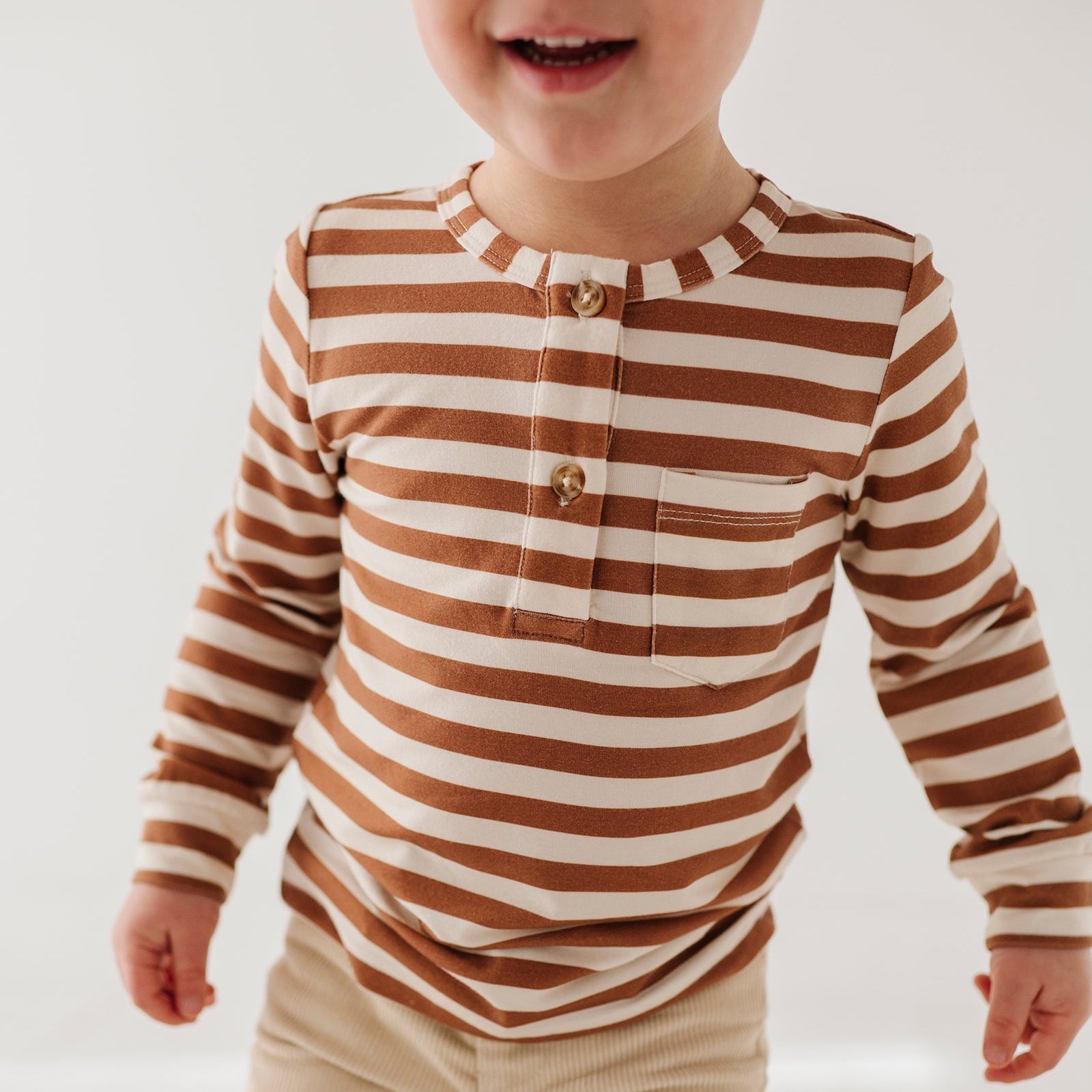 Child wearing a brown and white striped shirt with beige pants on a plain background