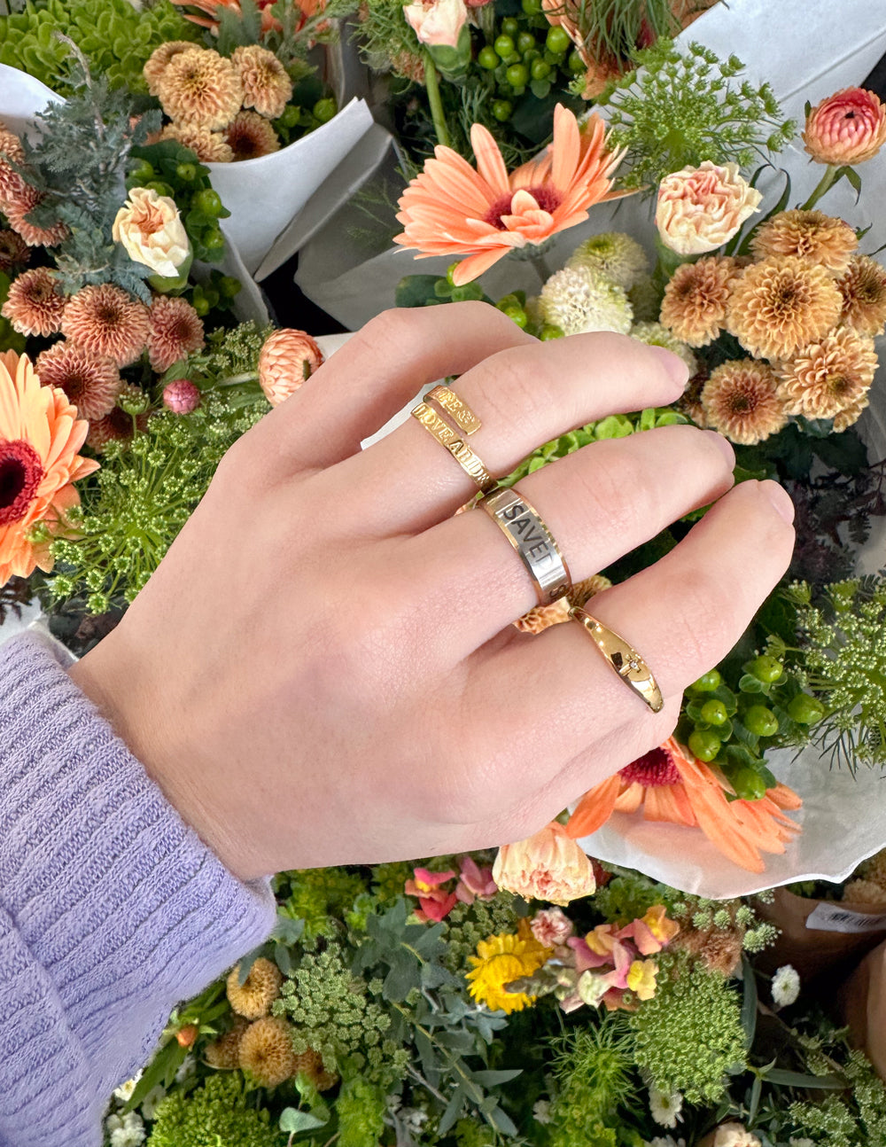 Hand wearing the Cross Gemstone Gold-Tone Ring with other rings beside colorful flowers