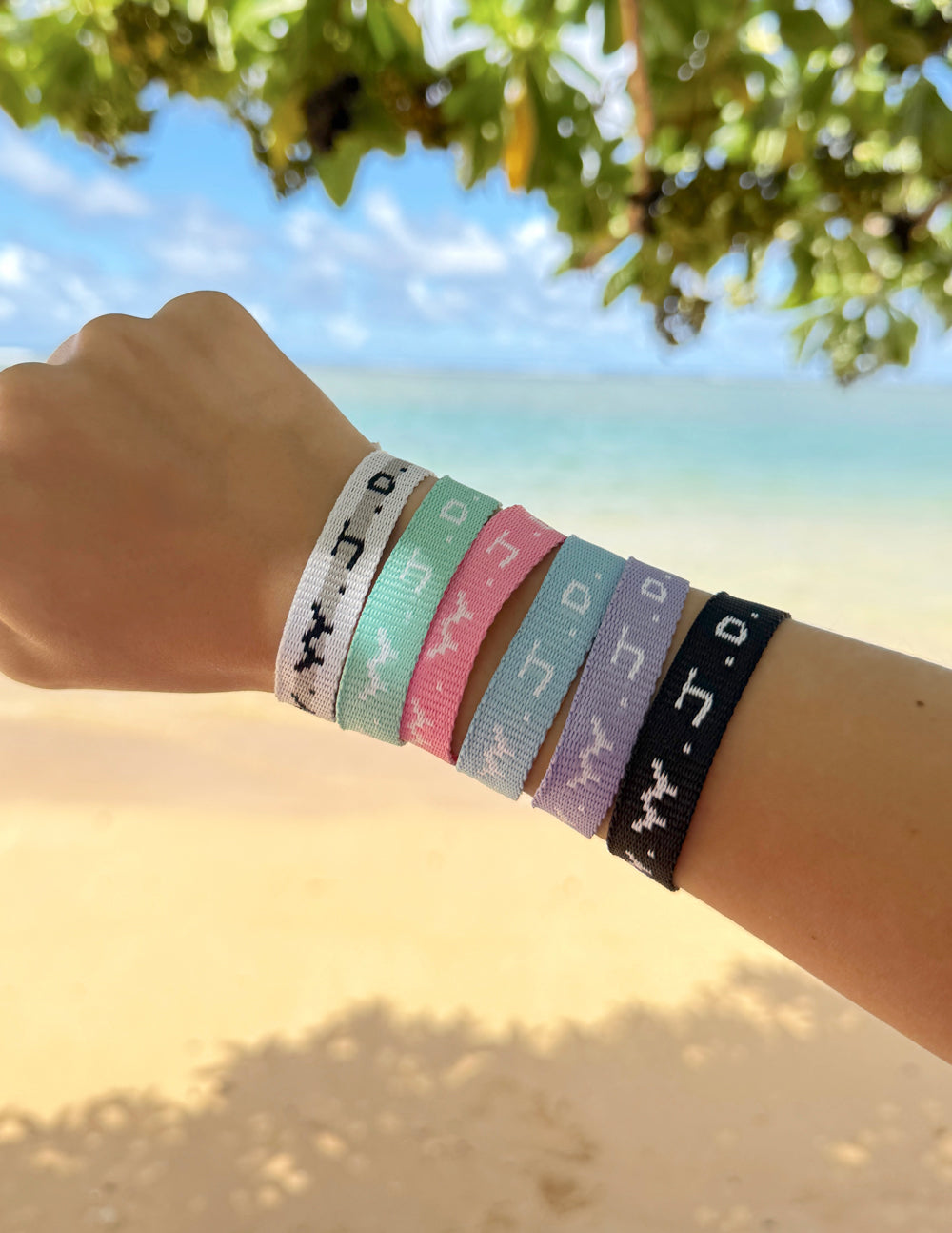Six assorted WWJD woven bracelets stacked on a wrist beside a sunny beach and turquoise water.