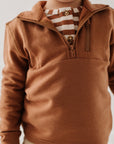 Child wearing a brown pullover with a striped shirt underneath on a plain background