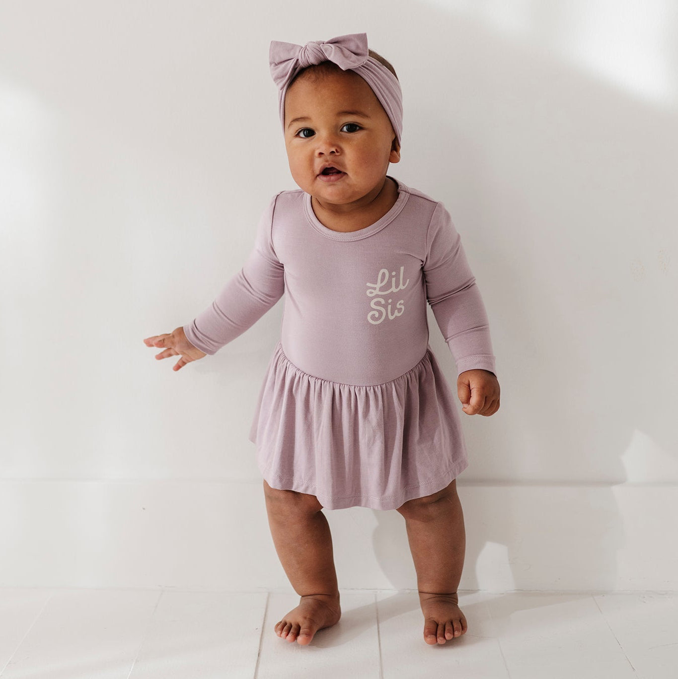 Baby in a pink outfit with a bow standing on a white floor.