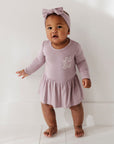 Baby in a pink outfit with a bow standing on a white floor.