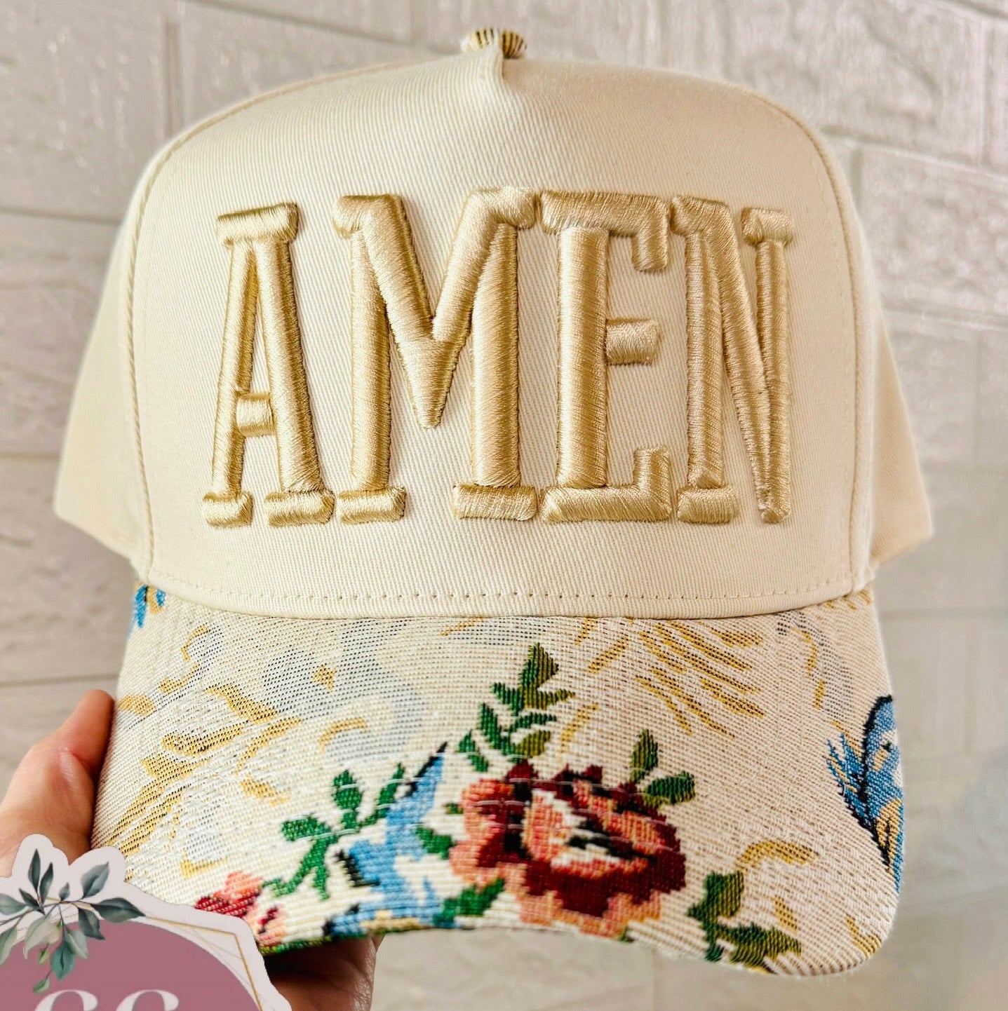 Cap with 'AMEN' embroidery and floral design, held against a light background