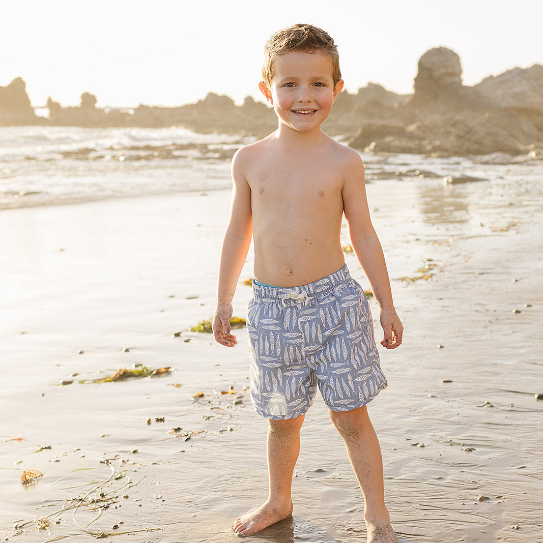 Young boy standing on a beach wearing patterned shorts.