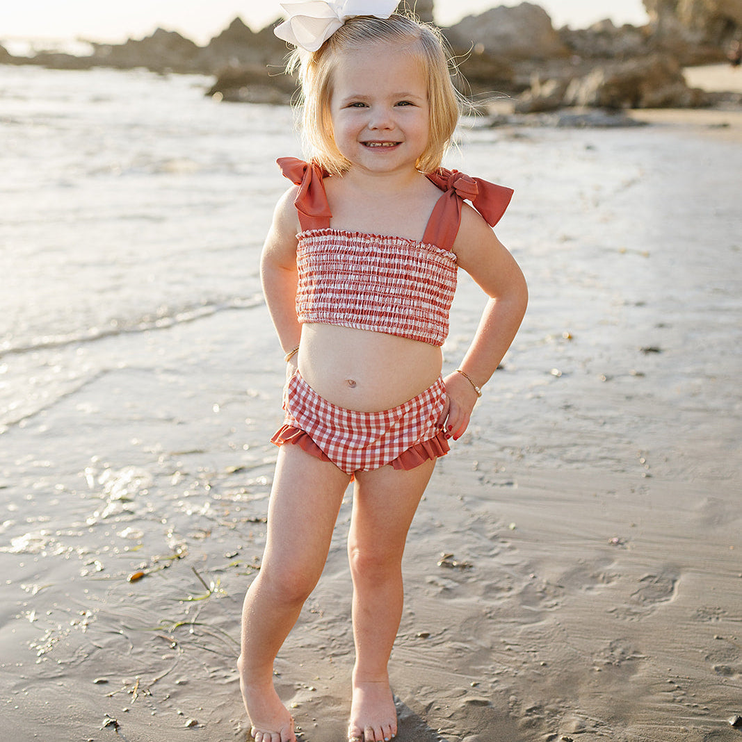 Young girl in a red checkered swimsuit standing on a sandy beach with rocks and water in the background.