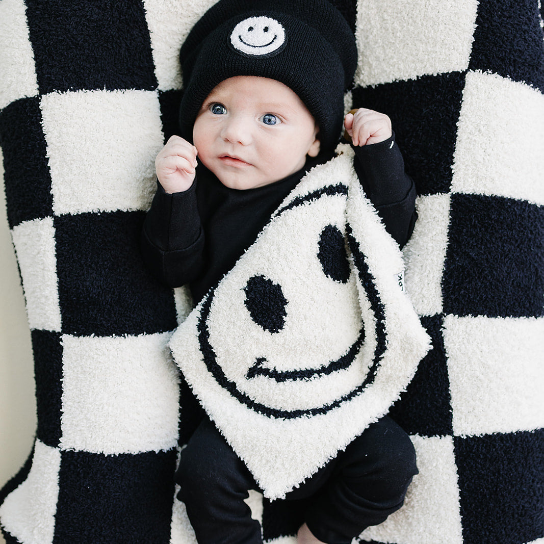 Baby wearing a black outfit with smiley face design, lying on a black and white checkered blanket.