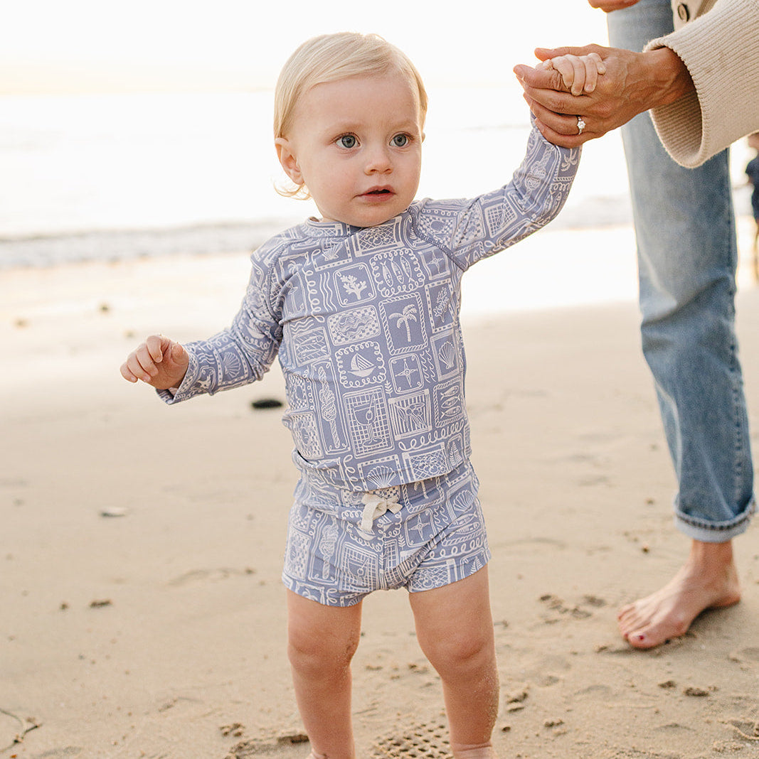 Baby in a patterned outfit standing on a beach with an adult's hand holding their other hand.