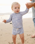 Baby in a patterned outfit standing on a beach with an adult's hand holding their other hand.