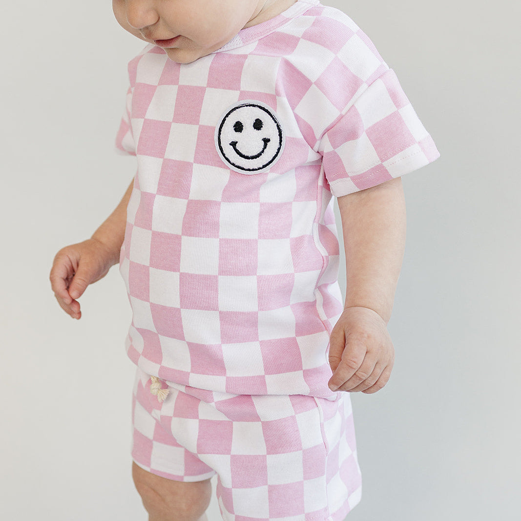 Baby wearing a pink and white checkered outfit with a smiley face design on a plain background