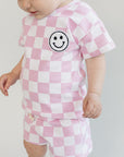 Baby wearing a pink and white checkered outfit with a smiley face design on a plain background
