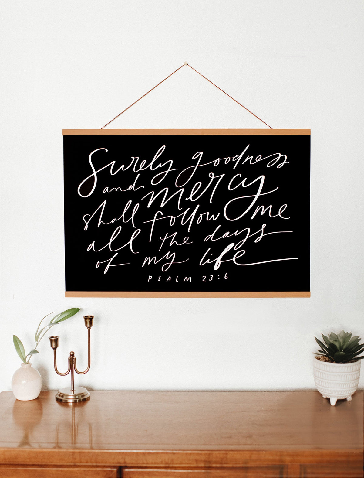 Surely Goodness Psalm 23:6 black wall art with white script and wooden hanging bars displayed above a console