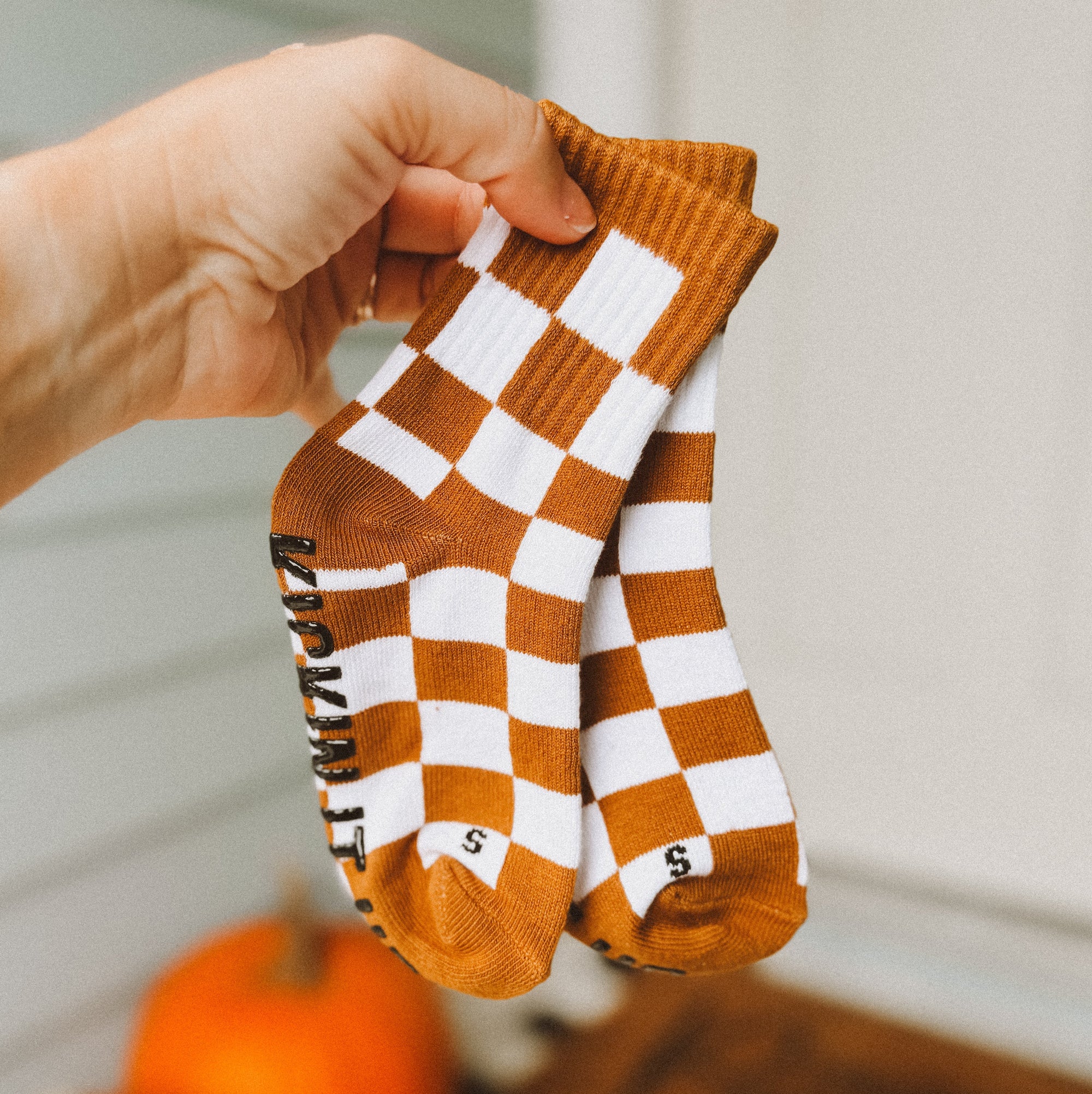 Checkered socks with brown and white pattern held by a hand, with pumpkins in the background.