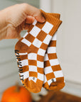 Checkered socks with brown and white pattern held by a hand, with pumpkins in the background.