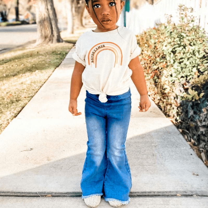 Child wearing a white t-shirt with a rainbow design and blue jeans on a sidewalk.