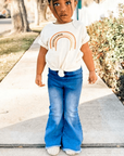 Child wearing a white t-shirt with a rainbow design and blue jeans on a sidewalk.