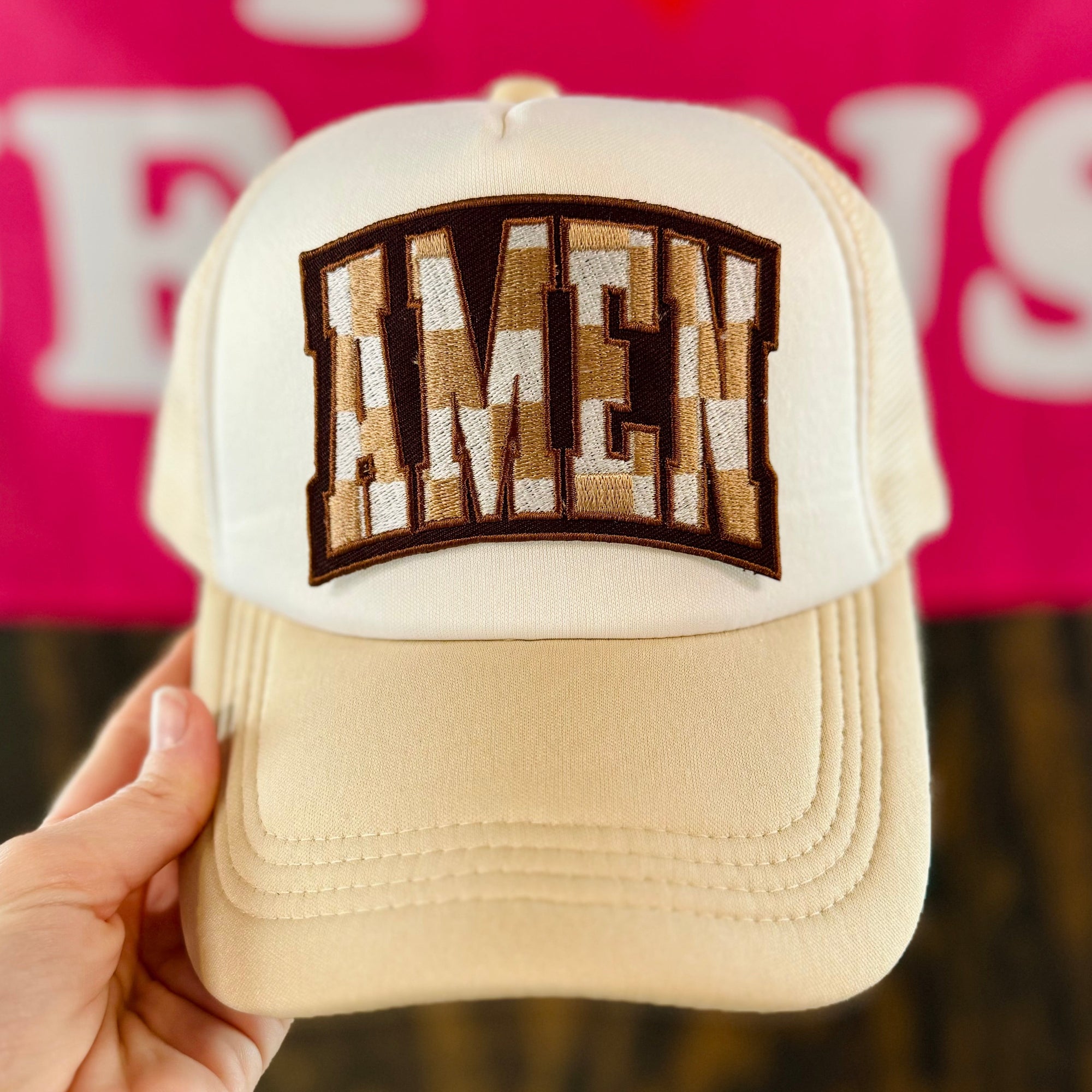 Beige cap with 'Amen' patch held by a hand against a pink background with text.