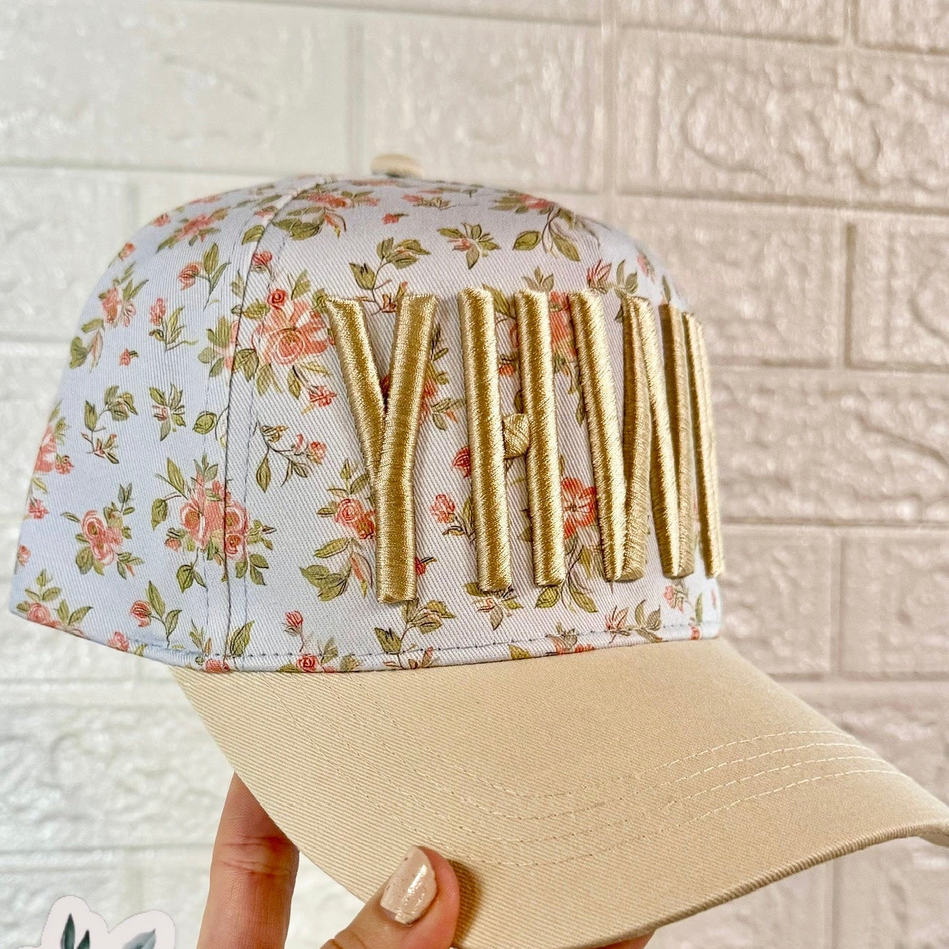 Floral-patterned cap with gold letters held by a hand against a tiled wall.